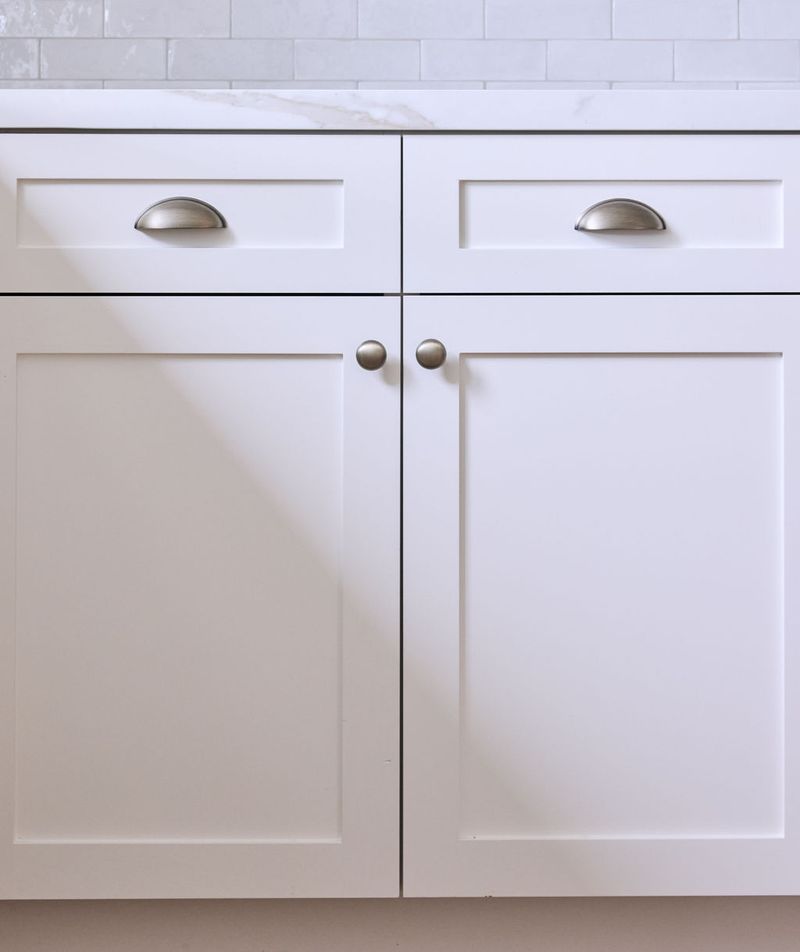 Close-up of white shaker-style base cabinets featuring two drawers with cup pulls and two lower cabinet doors with round knobs, topped with a marble countertop and white subway tile backsplash.