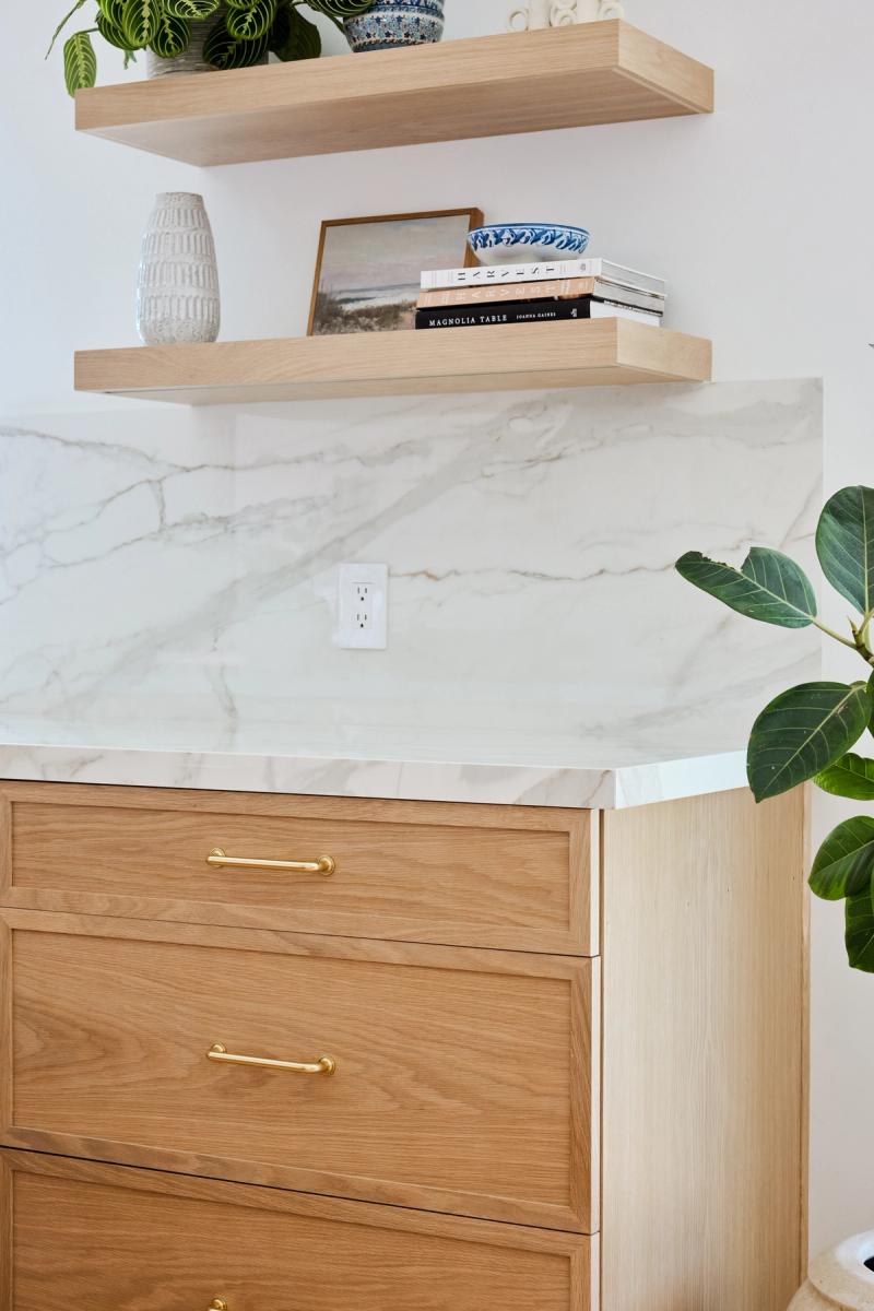 A light wood kitchen cabinet with brass bar pulls topped with a white marble countertop and matching slab backsplash, with two floating white oak shelves above holding books, a vase, and decorative objects.