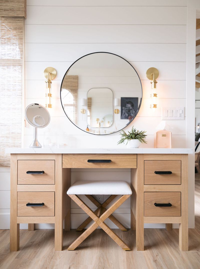 A custom white oak makeup vanity with a white marble countertop, black hardware, and five drawers flanking a center knee space. A round black-framed mirror hangs above on a shiplap wall, flanked by brass sconces, with an X-base upholstered stool tucked beneath.