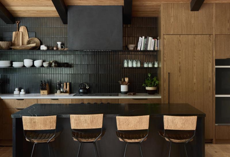 A modern kitchen featuring dark green vertical tile backsplash, white oak cabinetry, a black stone island with woven bar stools, open shelving, and a matte black range hood.