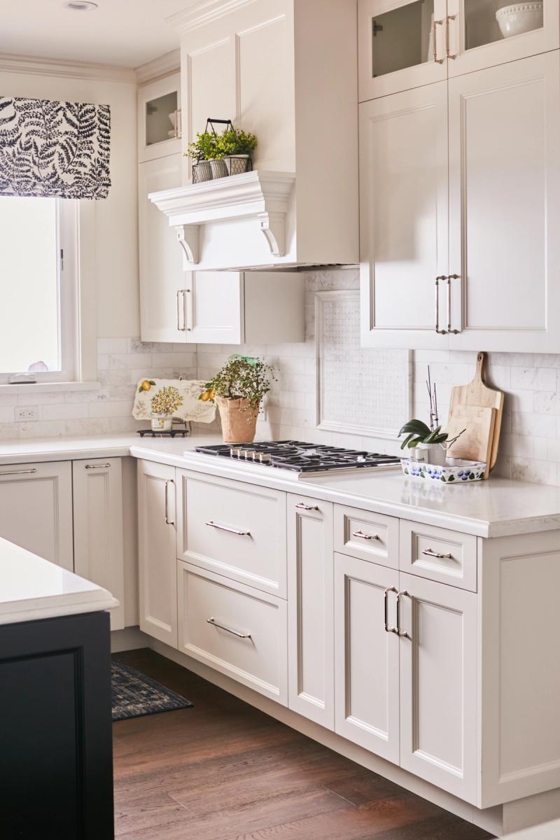 A bright, elegant kitchen featuring cream inset cabinetry with polished nickel hardware, a decorative white range hood, marble subway tile backsplash, white quartz countertops, and a built-in gas cooktop. A dark navy island is partially visible on the left.