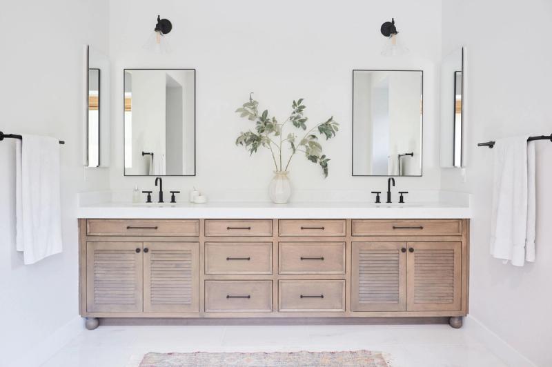 A wide double-sink bathroom vanity with natural wood cabinetry, louvered cabinet doors, matte black hardware, white quartz countertop, and two black-framed mirrors. A white vase with eucalyptus branches sits centered between the two sinks.