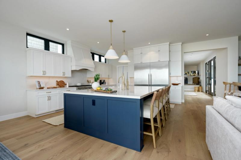 A bright, open-concept custom kitchen featuring floor-to-ceiling white shaker cabinets, a large navy blue island with white quartz countertop, gold pendant lights, and light hardwood flooring.