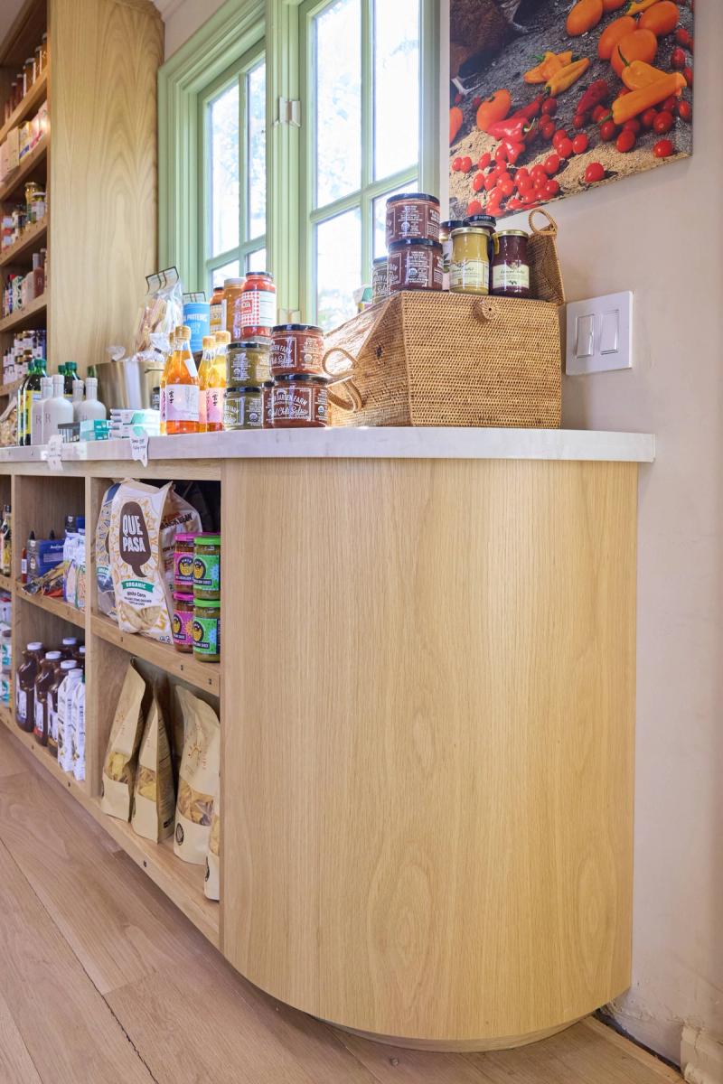 A curved natural oak display counter with a marble top sits in a bright specialty food shop, flanked by open wood shelving stocked with pantry goods, jars, and snacks.