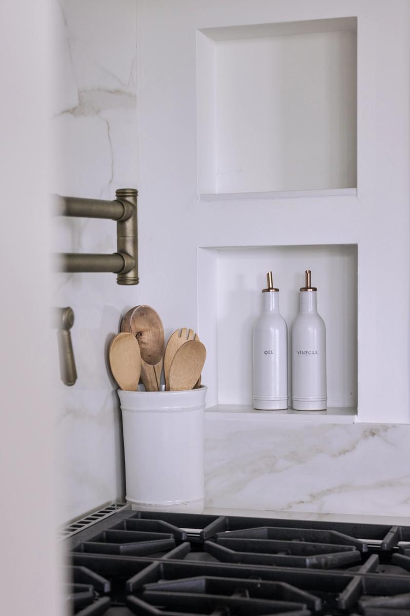 A close-up of a white kitchen backsplash area featuring built-in wall niches holding white ceramic oil and vinegar bottles, a utensil crock with wooden spoons, and a brass pot filler mounted on marble-look tile above a gas range.