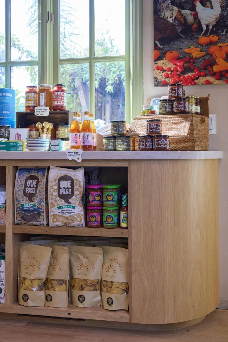 A curved light oak retail display counter with open shelving holding tortilla chips, organic snacks, and sauces, set in a bright shop with a large window.