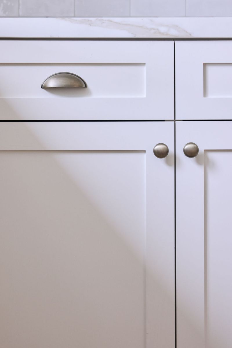 A close-up of white shaker-style base cabinets featuring a brushed nickel cup pull on a drawer and round knobs on double cabinet doors, beneath a marble countertop.