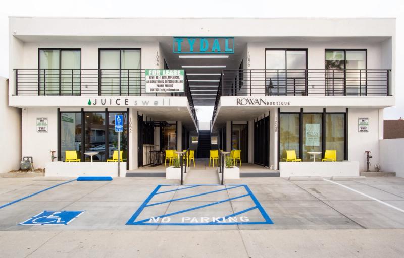 A modern two-story white commercial building in Ventura, California featuring ground-floor retail spaces for Juice Swell and Rowan Boutique, with yellow outdoor furniture, a central staircase, and upper-floor units for lease under the Tydal sign.