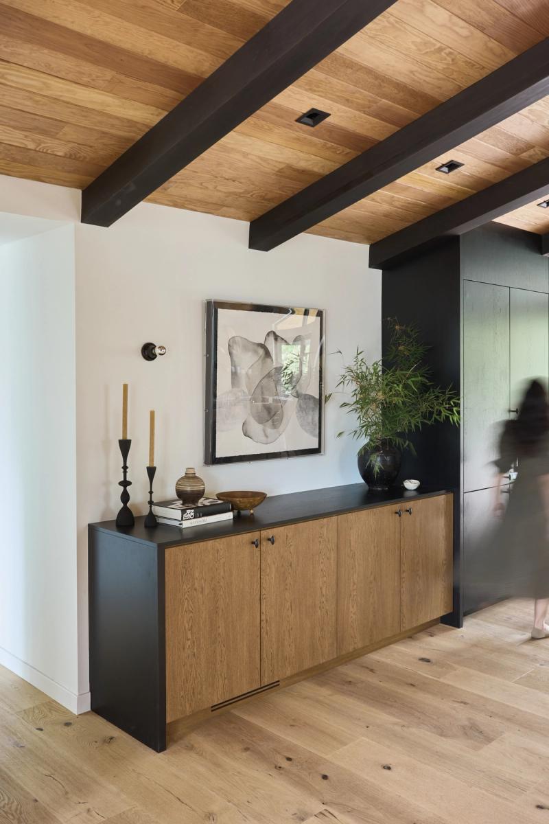 A custom built-in console cabinet with warm oak panel doors and a matte black frame, styled with candle holders, books, and a large potted plant beneath an abstract artwork, set in a room with wood-beamed ceilings and light oak flooring.