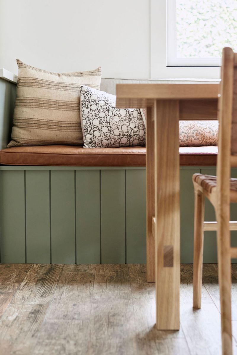A built-in banquette bench with sage green vertical shiplap paneling and a cognac leather cushion, styled with linen and block-print throw pillows, paired with a natural wood dining table and chair.