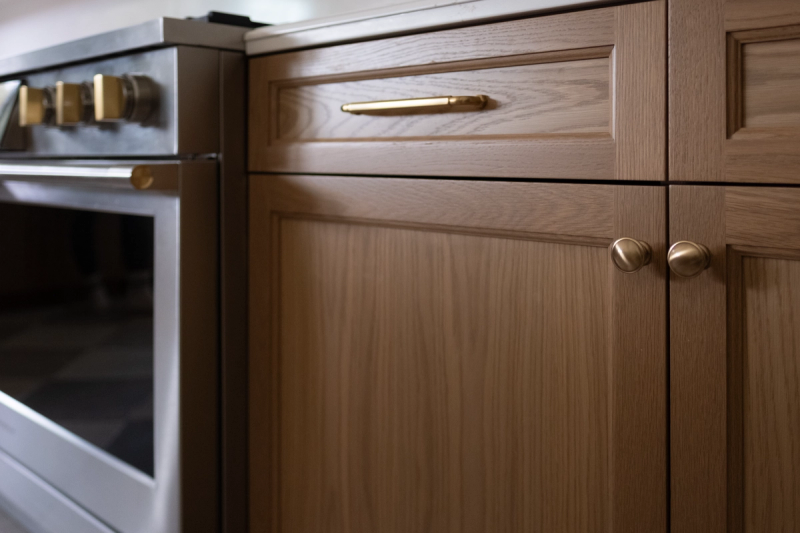 Close-up view of warm white oak kitchen base cabinets featuring inset panel doors and drawers with brushed brass pulls and knobs, alongside a stainless steel range with gold accents.