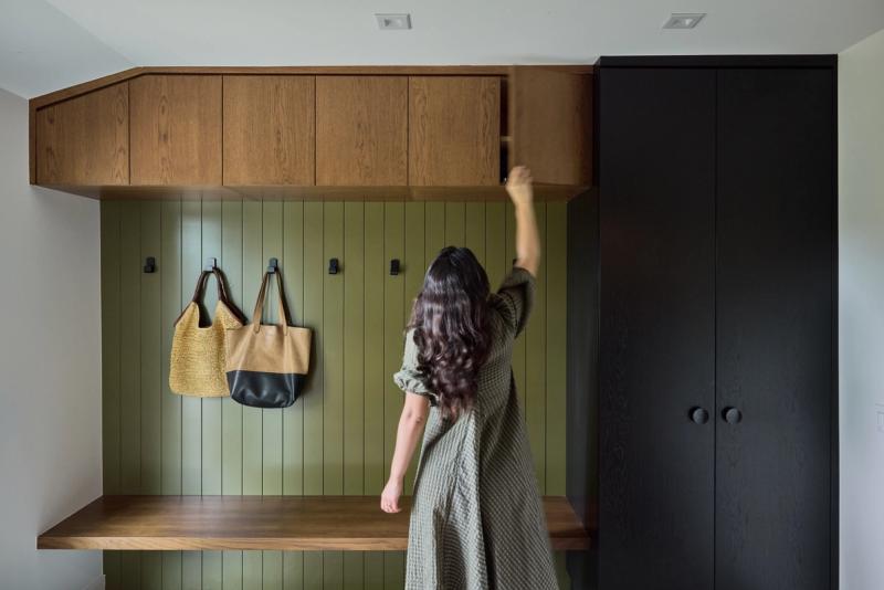 A woman in a green dress reaches up to open an upper wood cabinet in a custom mudroom featuring olive green shiplap walls, black hooks, a floating wood bench, and a tall black wardrobe cabinet.