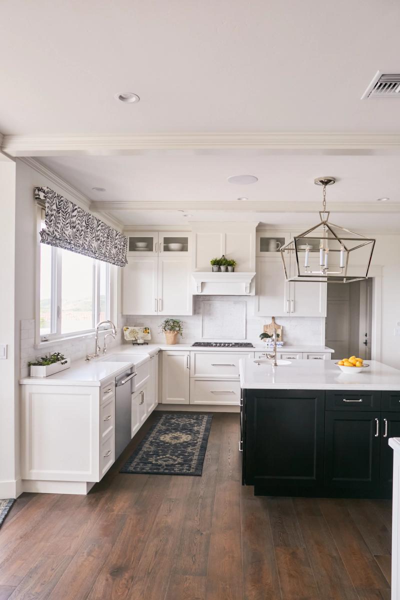 A bright, spacious custom kitchen featuring white shaker-style perimeter cabinets with chrome hardware, a dark navy-black island with white quartz countertops, hardwood floors, and a geometric lantern pendant light.