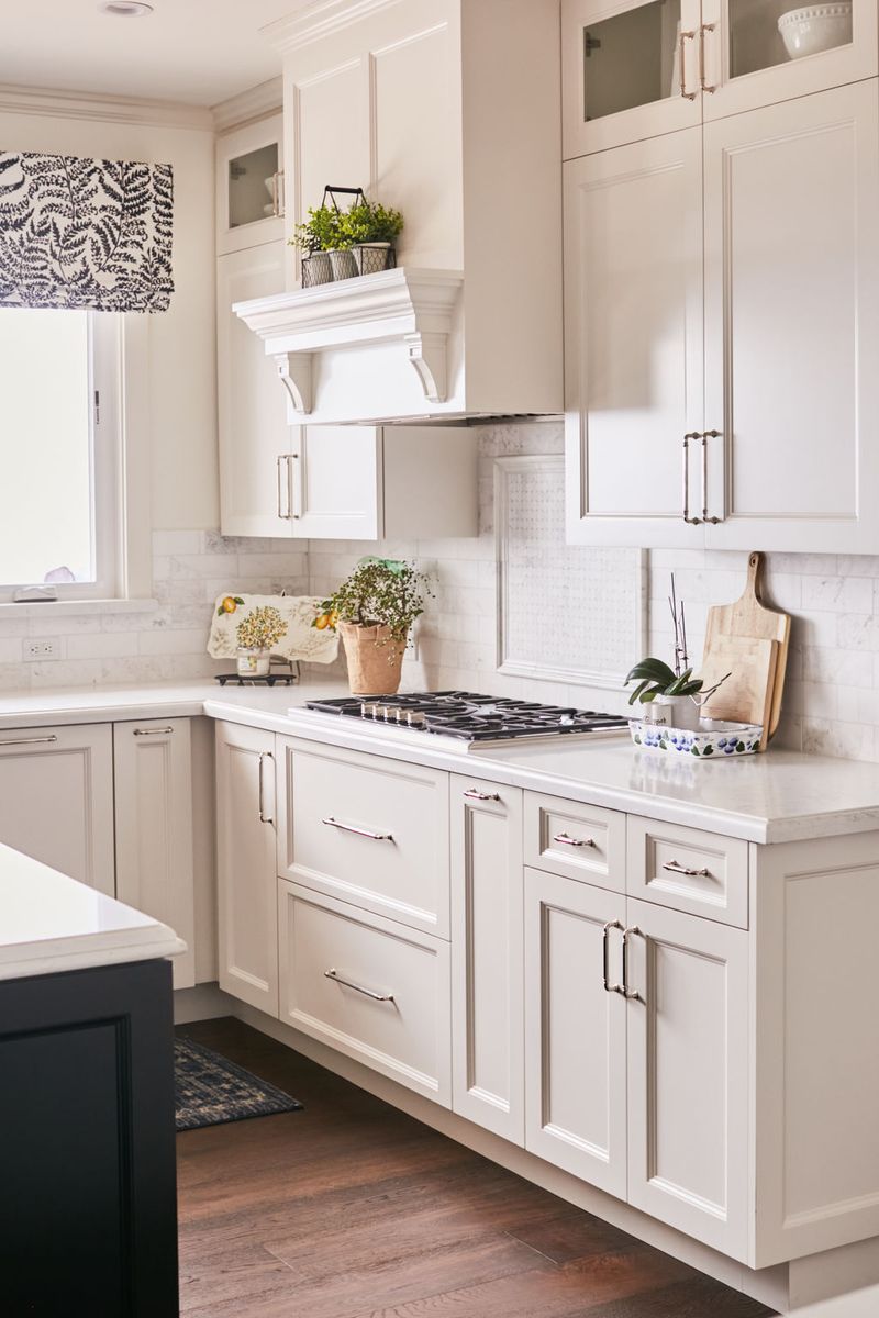 A bright, elegant kitchen featuring cream inset cabinetry with polished nickel hardware, a decorative white range hood, marble subway tile backsplash, white quartz countertops, and a built-in gas cooktop. A dark navy island is partially visible on the left.
