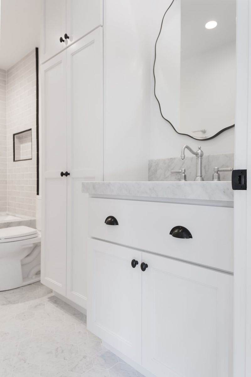 A bright white bathroom featuring custom shaker-style cabinetry with black cup pulls and knobs, a marble countertop, silver bridge faucet, wavy black-framed mirror, and marble tile flooring.