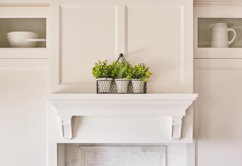 A bright white fireplace mantel with decorative corbels topped with a wire basket of green plants, flanked by glass-front built-in cabinetry displaying white dishware.