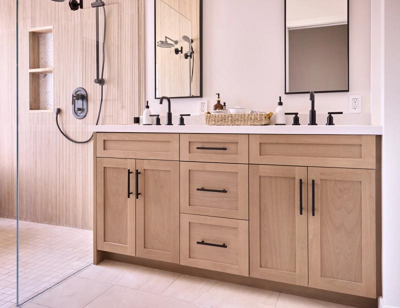 A modern bathroom featuring a wide double-sink vanity with light oak cabinetry, white countertop, matte black faucets and hardware, two framed mirrors, and an adjacent walk-in shower with fluted tile walls.