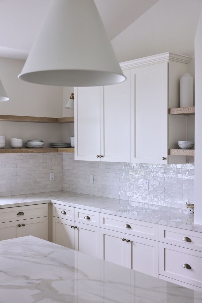A bright, elegant kitchen featuring cream shaker-style cabinetry with brushed nickel hardware, white marble countertops, iridescent subway tile backsplash, and open wood shelving in the corner.