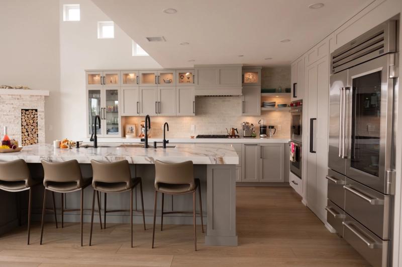 A spacious custom kitchen featuring gray shaker-style cabinetry, a large marble waterfall island with leather bar stools, stainless steel Sub-Zero appliances, and warm wood flooring with high vaulted ceilings.