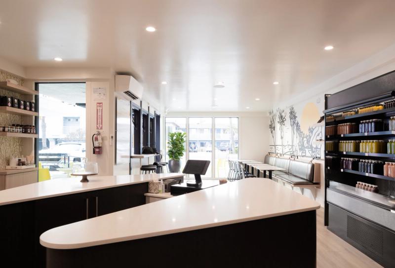 A bright, modern commercial interior featuring a curved white quartz service counter with dark cabinetry, open retail shelving stocked with bottled juices, and a seating area along the back wall with a botanical mural.