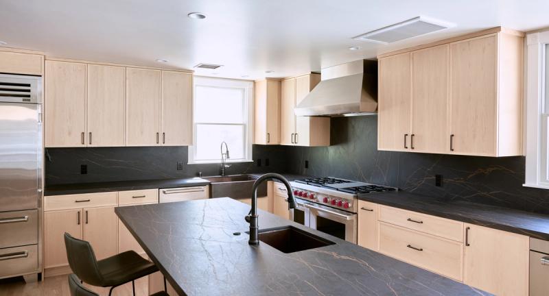 A spacious modern kitchen featuring light maple shaker-style cabinetry, dark soapstone countertops and backsplash, a large center island with an undermount sink, and a professional-grade stainless steel range with a wall-mounted hood.