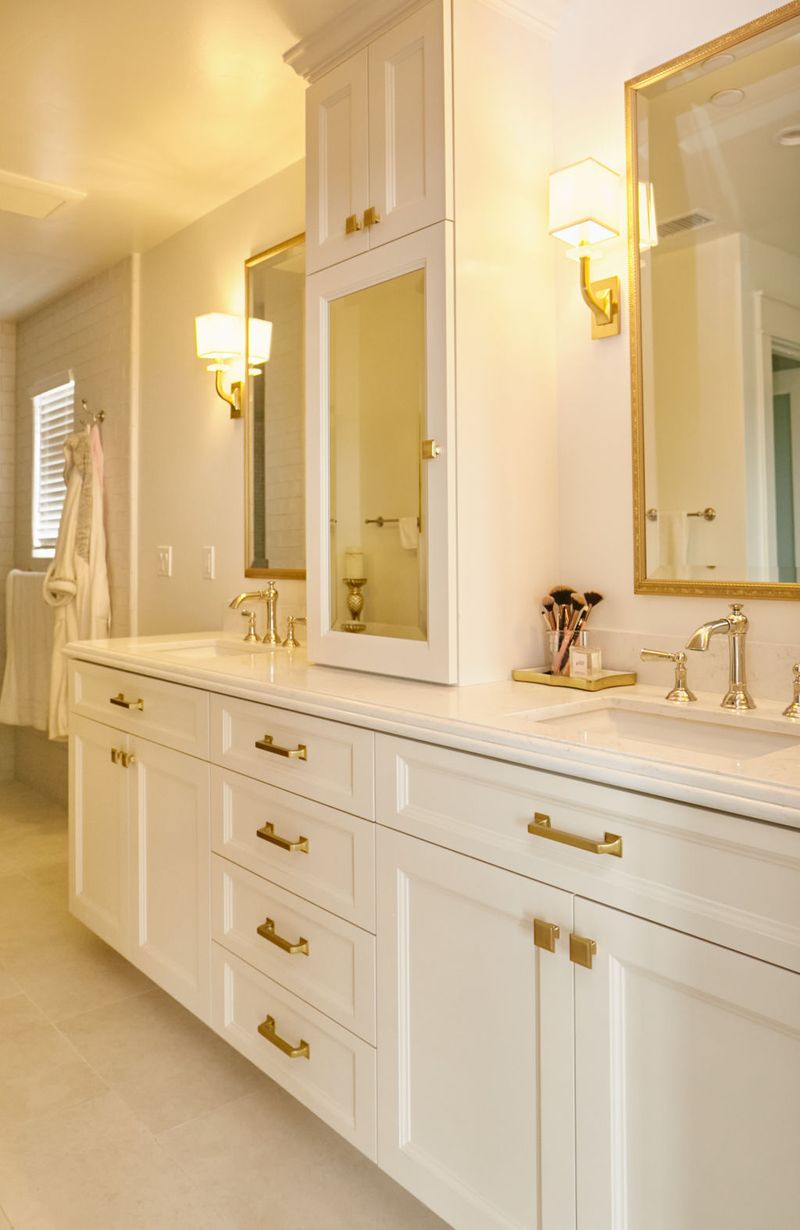A bright, elegant master bathroom featuring a white double vanity with gold hardware, framed mirrors, wall sconces, and a center tower cabinet with a mirrored door.