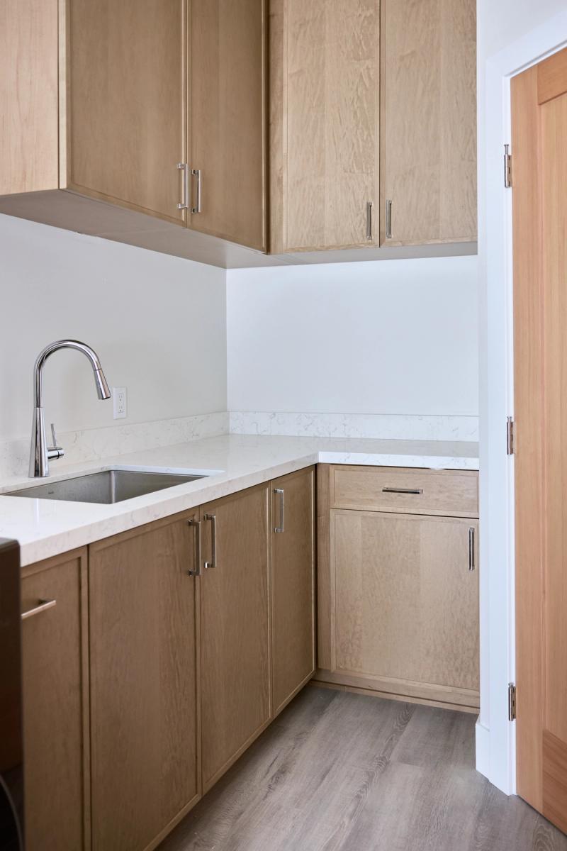 A modern kitchen corner featuring light oak flat-panel cabinetry, quartz countertops, an undermount stainless steel sink with a chrome pull-down faucet, and upper cabinets with bar-pull hardware.