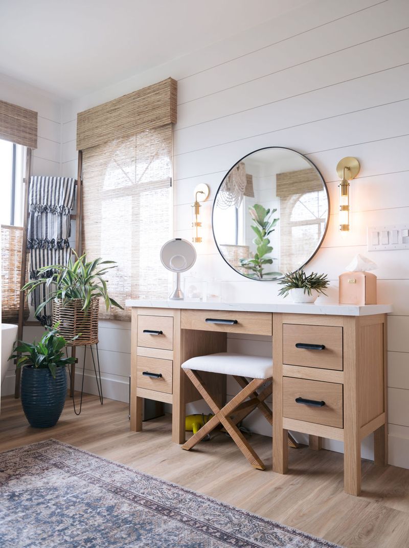 A bright, airy makeup vanity area featuring a light oak cabinet with black hardware and a white marble countertop, flanked by brass wall sconces and a round black-framed mirror, set against a white shiplap wall with natural woven window shades.