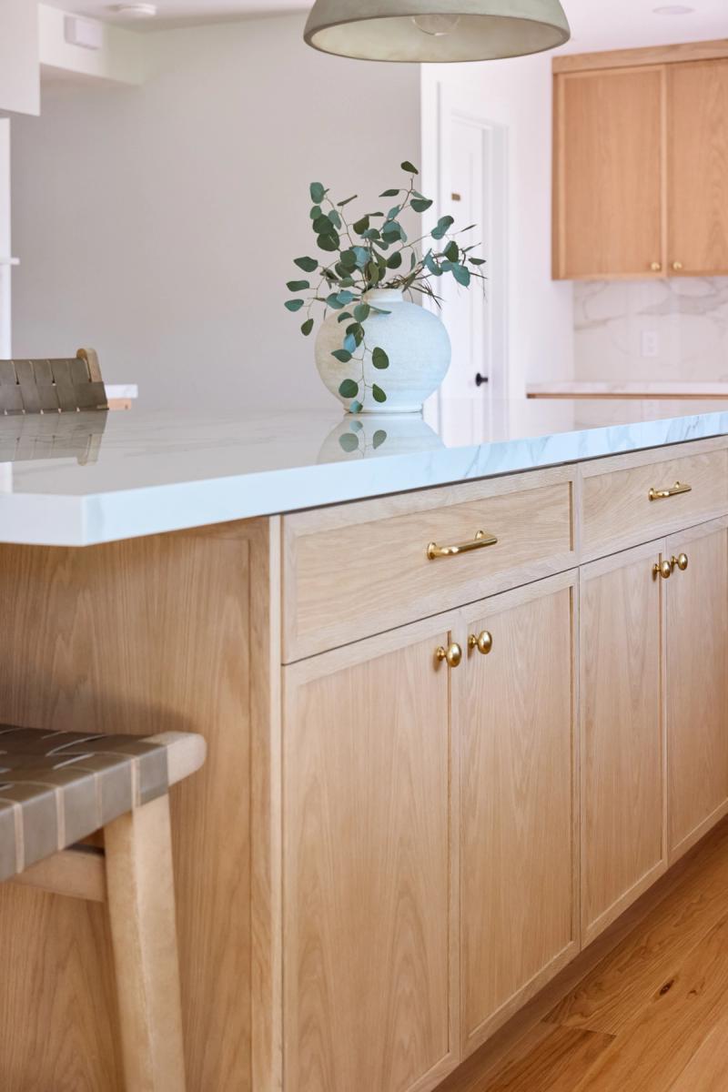 A large white oak kitchen island featuring brass cabinet pulls and knobs, topped with a white marble-look quartz countertop and decorated with a ceramic vase holding eucalyptus branches.