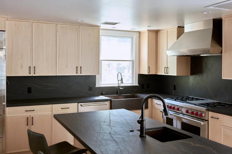 A modern kitchen featuring flat-panel light oak cabinetry, dark soapstone countertops and backsplash, a copper farmhouse sink, professional-grade range, and a large island with a second sink.