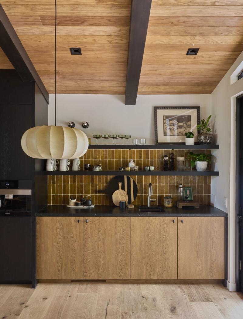 A modern kitchen bar area featuring natural oak lower cabinets, a dark countertop, olive-toned subway tile backsplash, open black floating shelves with glassware and plants, and a wood-paneled ceiling with dark beams.