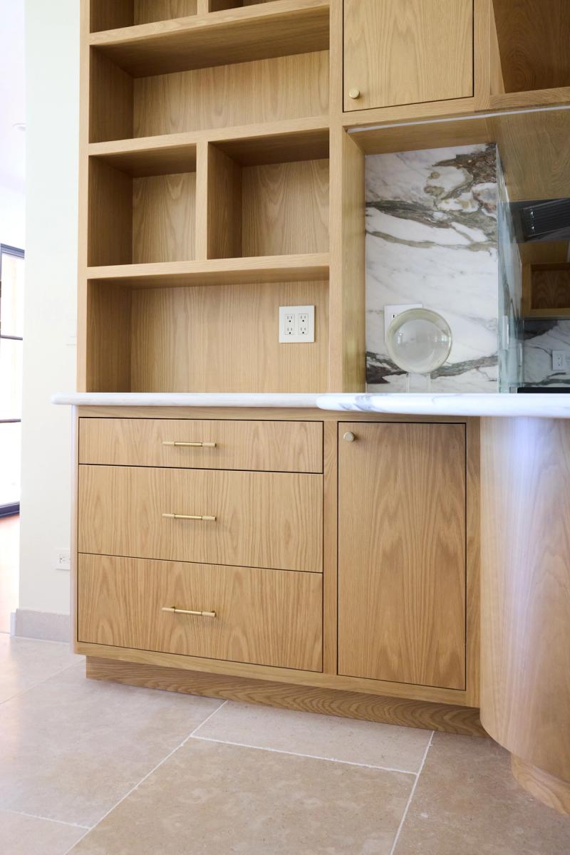 A custom natural oak kitchen cabinet unit featuring three drawers with brass bar pulls, an adjacent cabinet door with a round knob, open upper shelving cubbies, and a white marble countertop and backsplash.