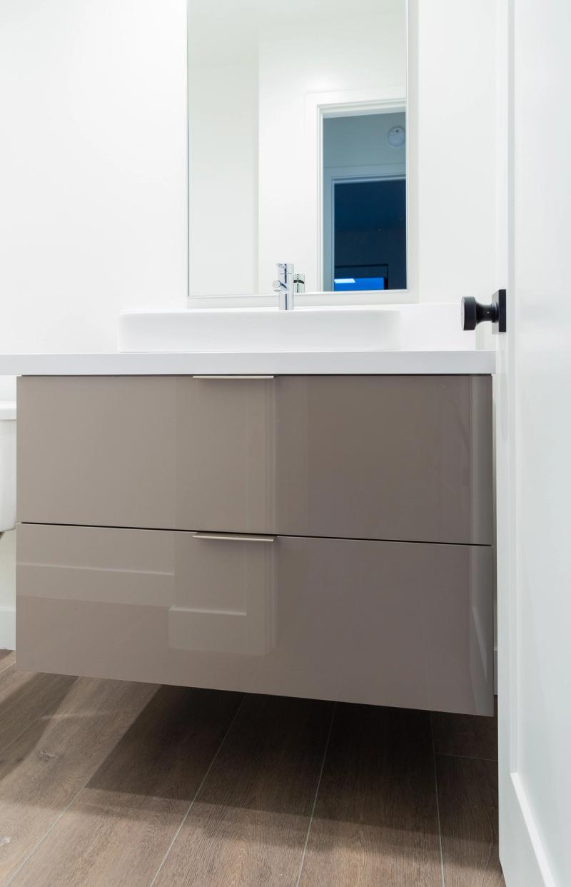A modern floating bathroom vanity with high-gloss taupe drawer fronts, white integrated sink, chrome faucet, and a frameless mirror above, set against white walls with wood-look tile flooring.