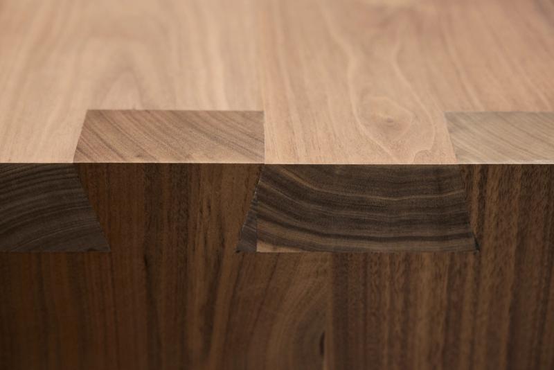 A close-up macro shot of a walnut wood joint where a tabletop or shelf meets a vertical panel, showcasing the natural grain contrast between lighter and darker wood tones.