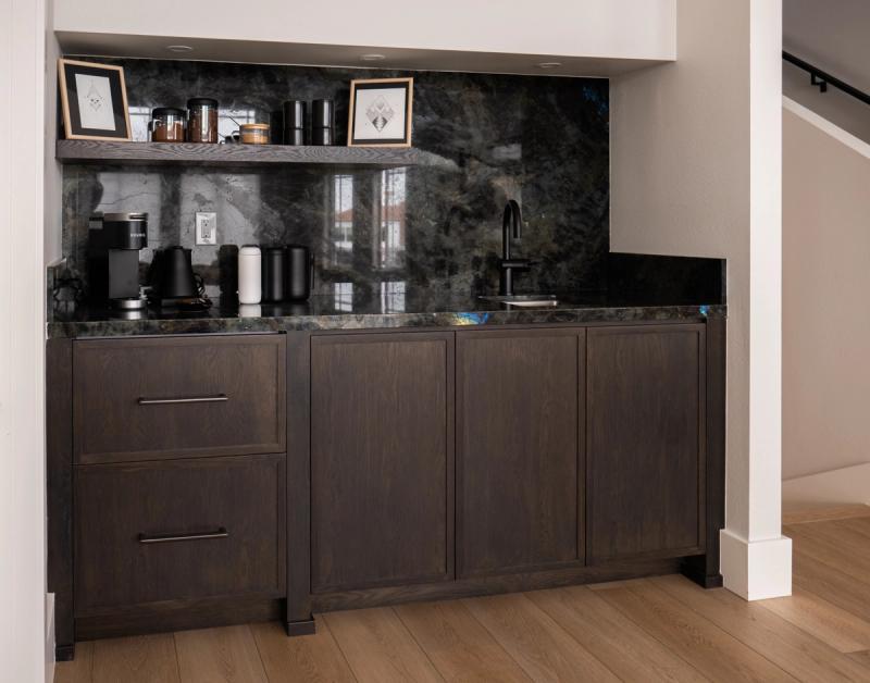 A custom dark oak coffee bar station with a dramatic black and green labradorite stone backsplash and countertop, featuring two drawers, cabinet doors, a small sink with matte black faucet, and an open shelf with coffee appliances and decorative items.