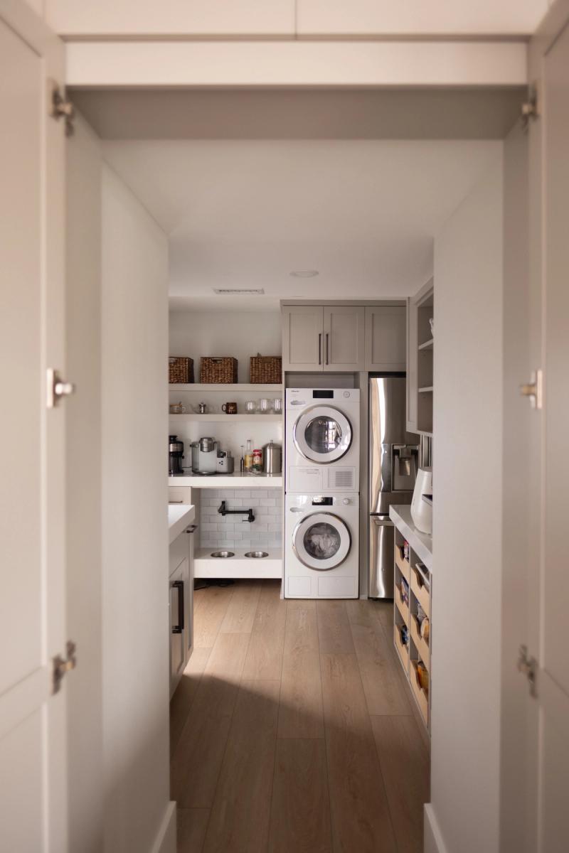 A well-organized butler's pantry featuring gray shaker cabinets, open shelving with wicker baskets and small appliances, a stacked washer and dryer unit, and pull-out storage drawers, viewed through open bifold doors.