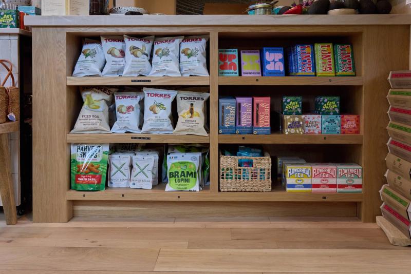 A light oak built-in shelving unit with three rows of shelves displaying packaged snacks, chocolates, and teas in a retail shop setting. The cabinetry features clean lines and a warm natural wood finish.