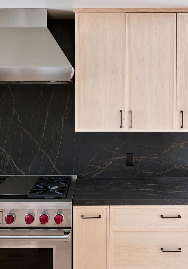 A modern kitchen featuring light maple flat-panel cabinets with black bar pulls, dramatic black marble-veined stone countertops and backsplash, and a professional stainless steel range with red knobs.