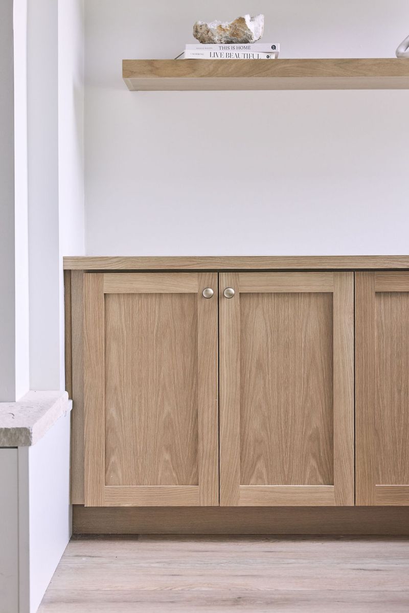 Light natural oak shaker-style base cabinets with brushed nickel knobs sit beneath a matching floating wood shelf styled with books and a raw stone, set against white walls with light hardwood floors.