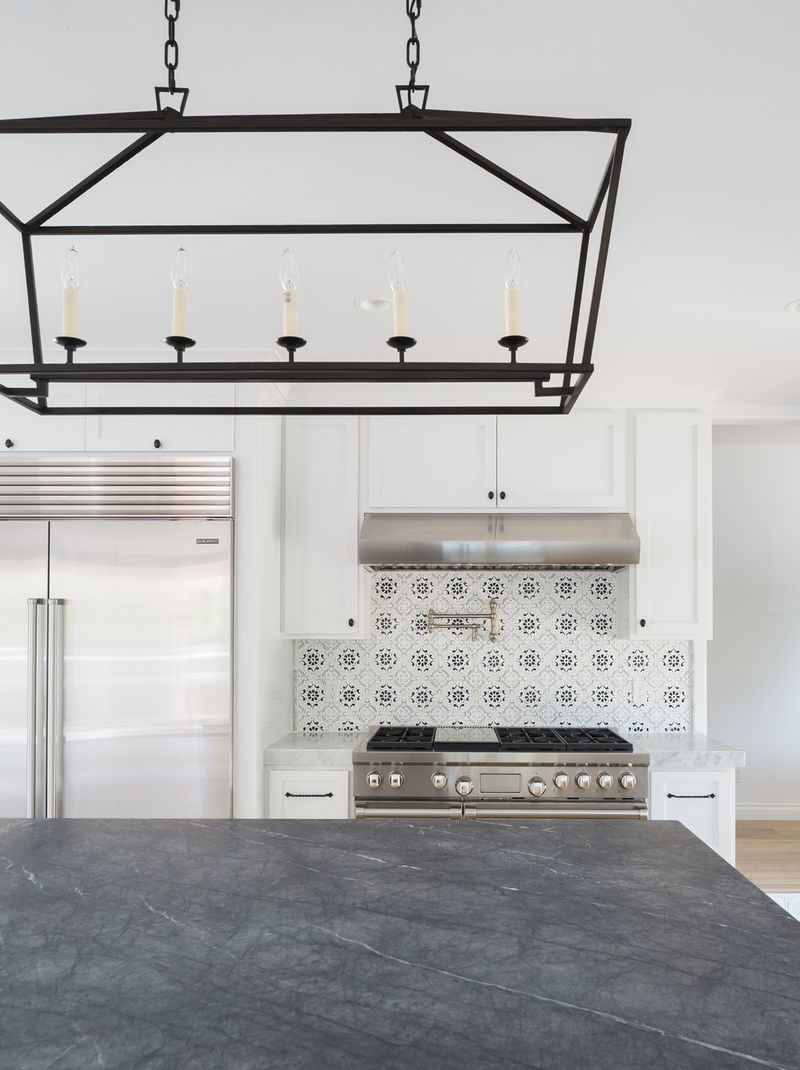A bright, high-end kitchen featuring white shaker cabinets, a stainless steel professional range with decorative tile backsplash, a large dark soapstone island, and a black iron linear chandelier overhead.