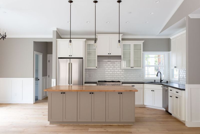 A bright, open kitchen featuring white upper cabinets, a greige island with butcher block countertop, stainless steel appliances, white subway tile backsplash, and three pendant lights over the island.