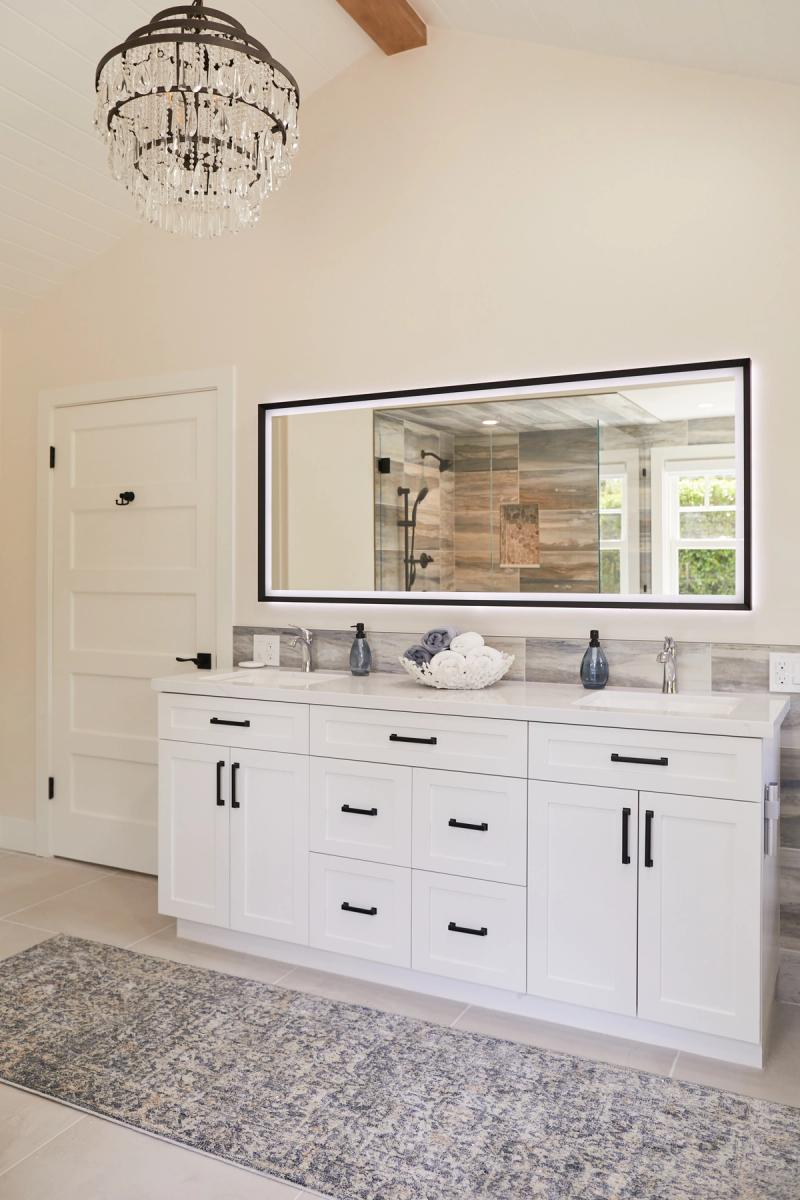 A bright, spacious master bathroom featuring a large white shaker-style double vanity with black hardware, a wide LED-backlit mirror, marble countertops, and an elegant crystal chandelier above a patterned area rug.