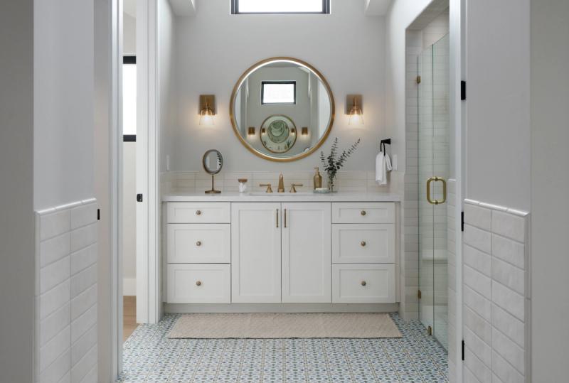 A bright, elegant bathroom featuring a white shaker-style vanity with gold hardware, a round gold-framed mirror, patterned floor tile, and a glass-enclosed shower visible to the right.
