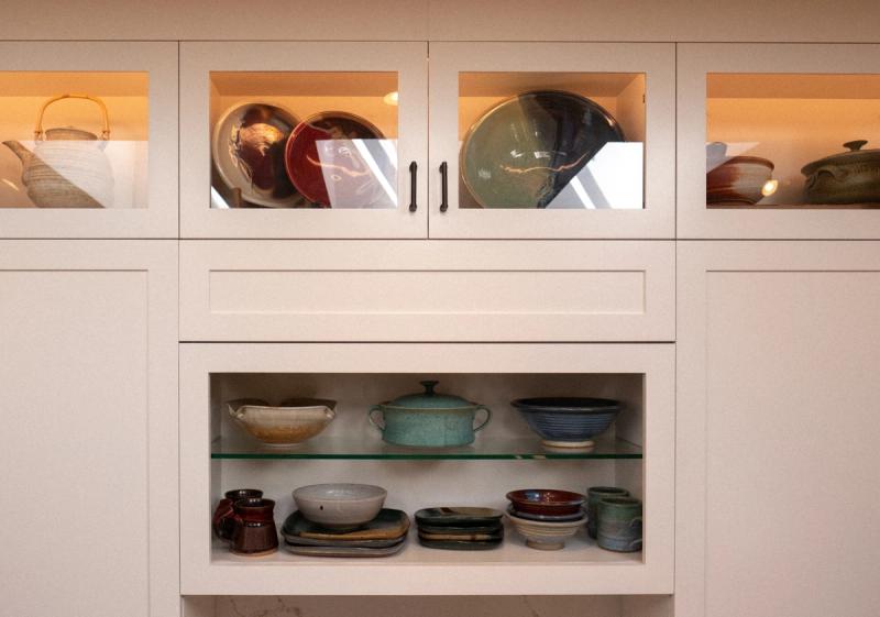 A cream-colored shaker-style built-in cabinet with glass-front upper doors displaying ceramic plates and a teapot, and open lower shelves holding an assortment of handmade pottery bowls, mugs, and plates.