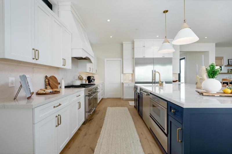 A bright, spacious custom kitchen featuring white shaker cabinets with brass hardware along the perimeter, a large navy blue island with white quartz countertops, stainless steel appliances, and warm wood flooring.