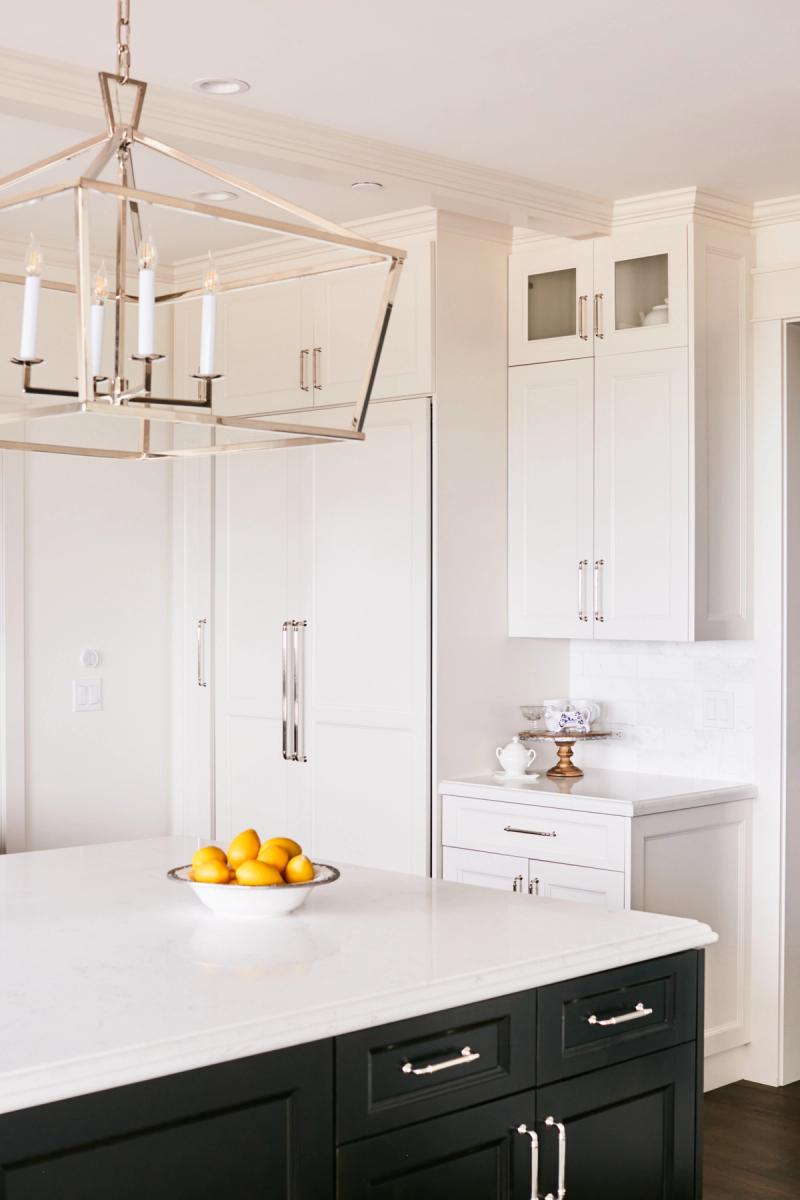 A bright, elegant kitchen featuring white shaker-style wall cabinets and a dark navy or black island with white quartz countertops, polished nickel hardware, a lantern-style chandelier, and a bowl of lemons on the island.