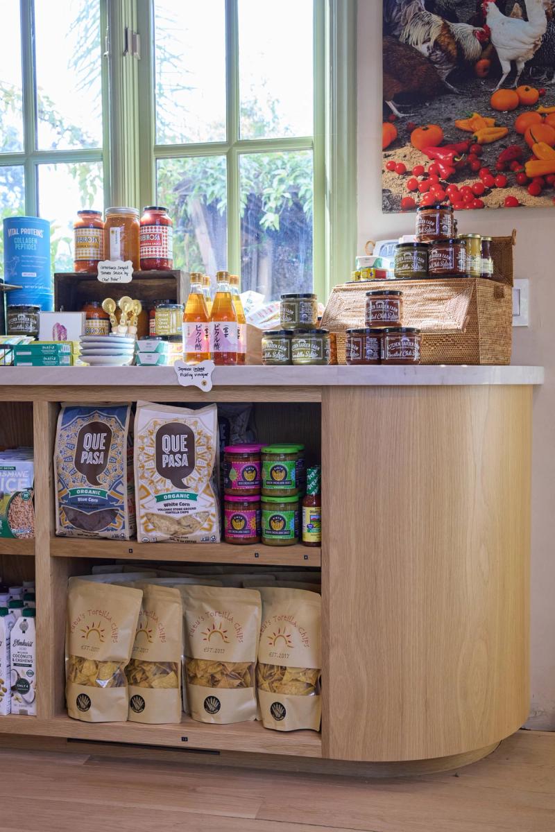 A curved light oak display cabinet with open shelving holds packaged snacks, sauces, and specialty food products in a bright retail or boutique shop setting with large windows.
