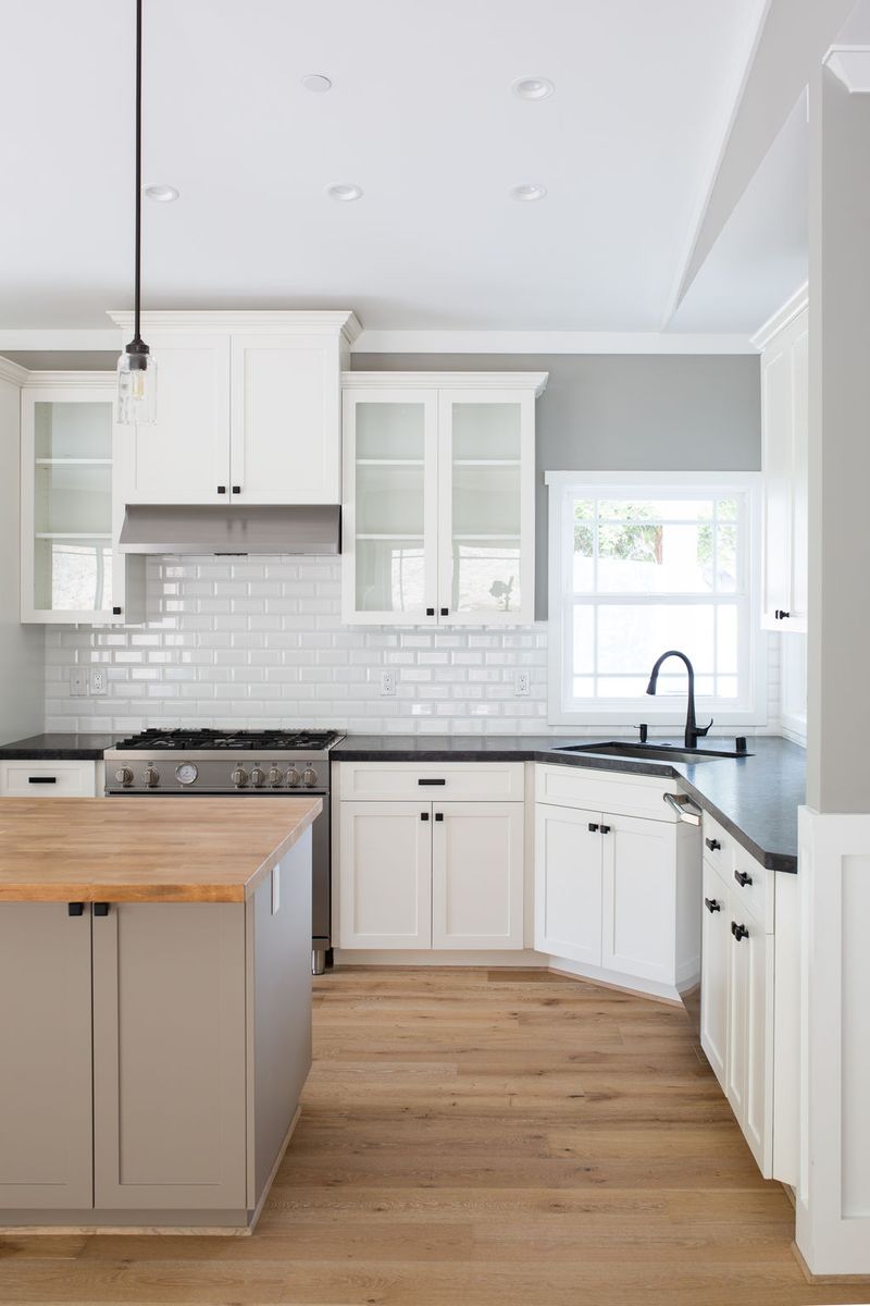 A bright, transitional kitchen featuring white shaker cabinets with black hardware, dark stone countertops, a white subway tile backsplash, stainless steel range, and a gray island with a butcher block top on light oak hardwood floors.