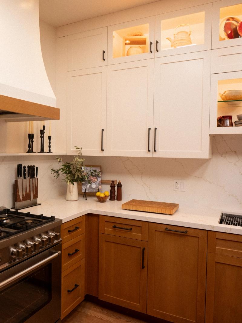 A custom kitchen featuring white shaker upper cabinets with glass accent panels and warm natural wood lower cabinets, paired with white marble-look countertops, black hardware, and a professional stainless steel range.