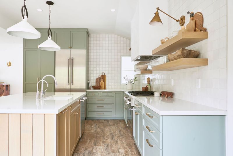 A bright, custom kitchen featuring sage green upper cabinets, light blue-green lower cabinets, a natural wood island, white quartz countertops, floating wood shelves, and white tile backsplash with brass hardware throughout.
